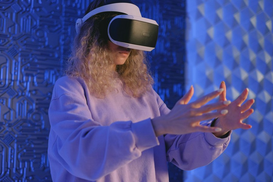 Woman with curly blonde hair wearing a VR headset, reaching out with both hands against a blue patterned wall