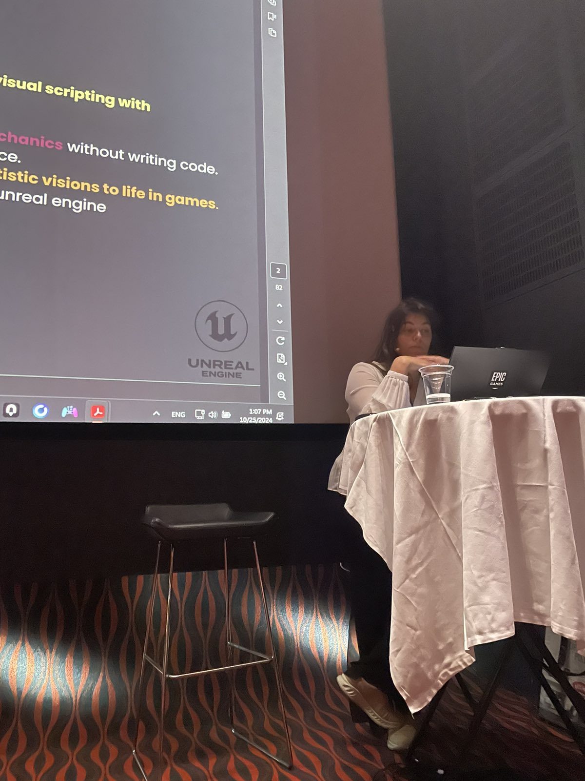 Elena Dimopoulou presenting an Unreal Engine visual scripting workshop at a laptop, slides projected behind her